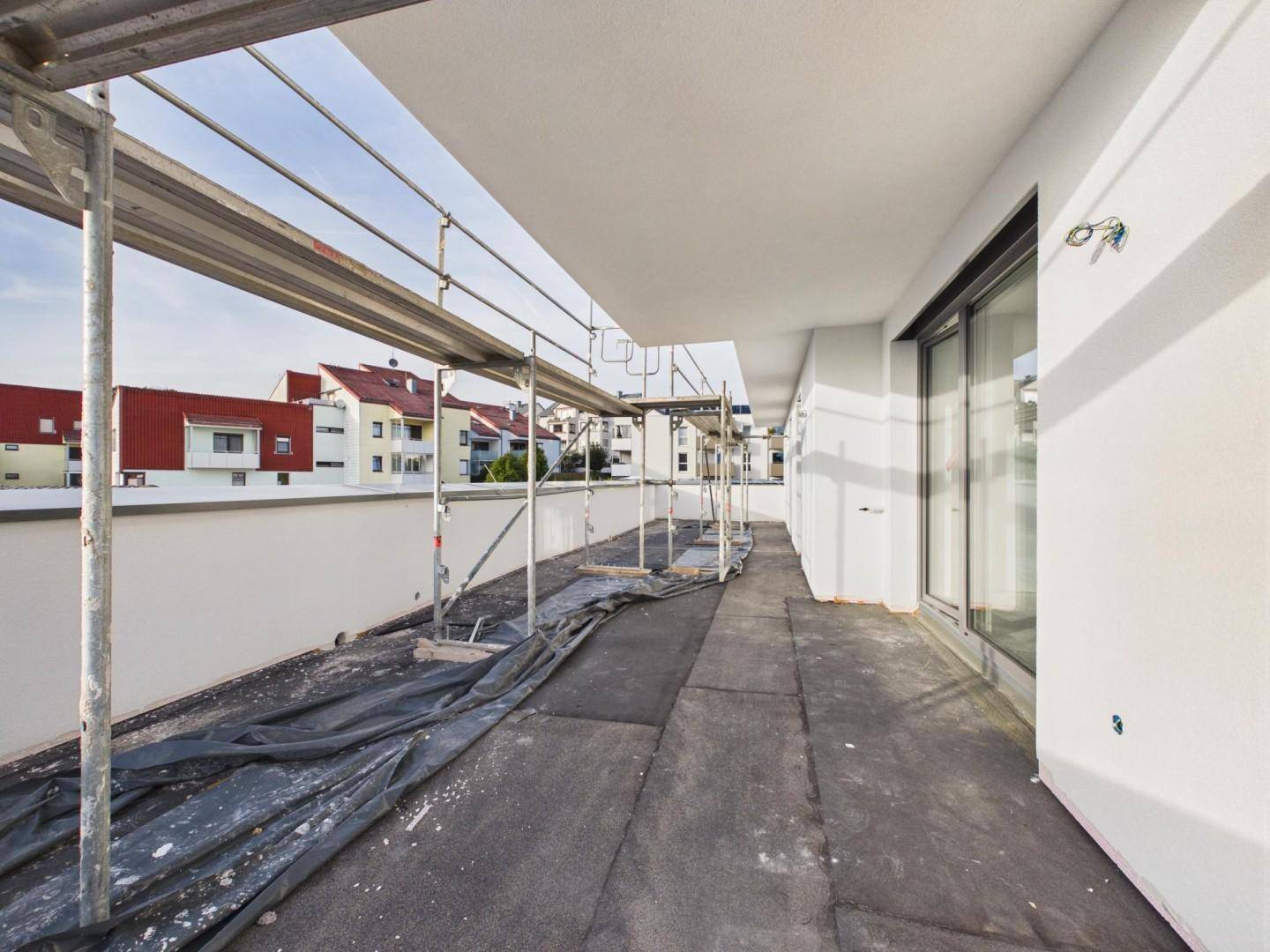 Langer Balkon eines Neubaus im Rohbauzustand mit Gerüst und Blick auf umliegende Gebäude.
