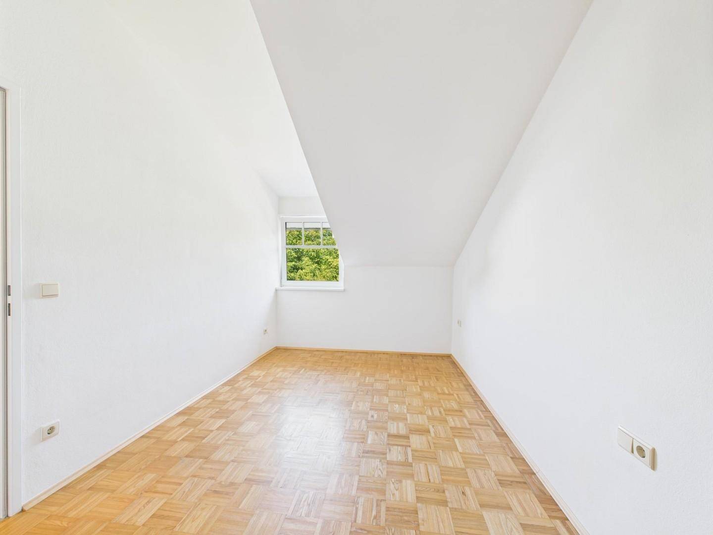 Geräumiges Zimmer mit Dachschräge, Parkettboden und Fenster mit Blick auf Bäume.
