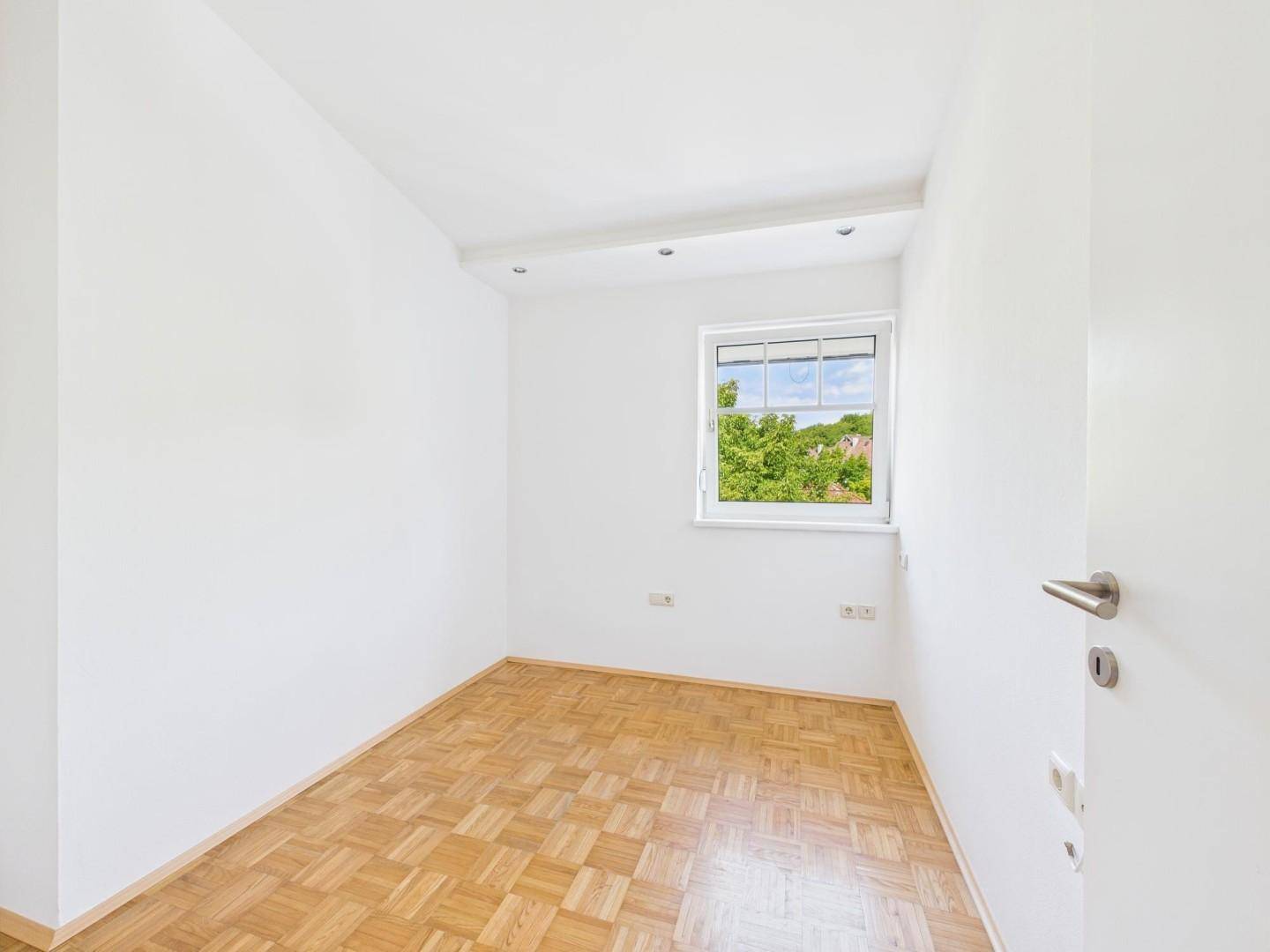 Helles Zimmer mit Parkettboden, weißen Wänden und einem Fenster mit Blick ins Grüne.