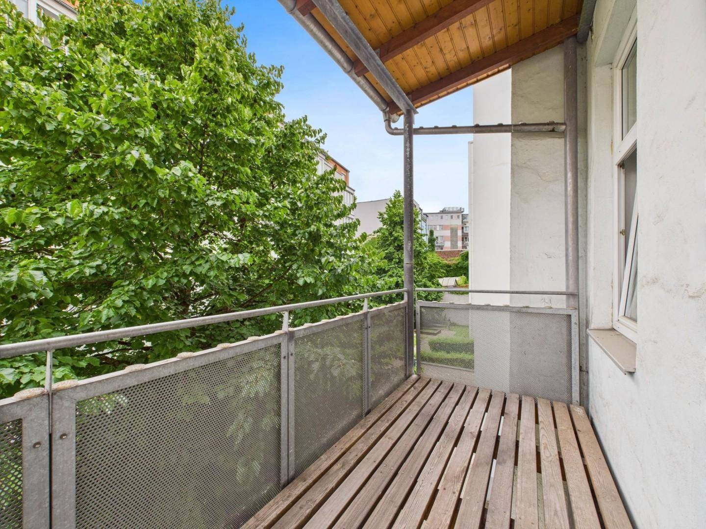 Kleiner Balkon mit Holzboden und Metallgeländer, umgeben von grünen Bäumen.