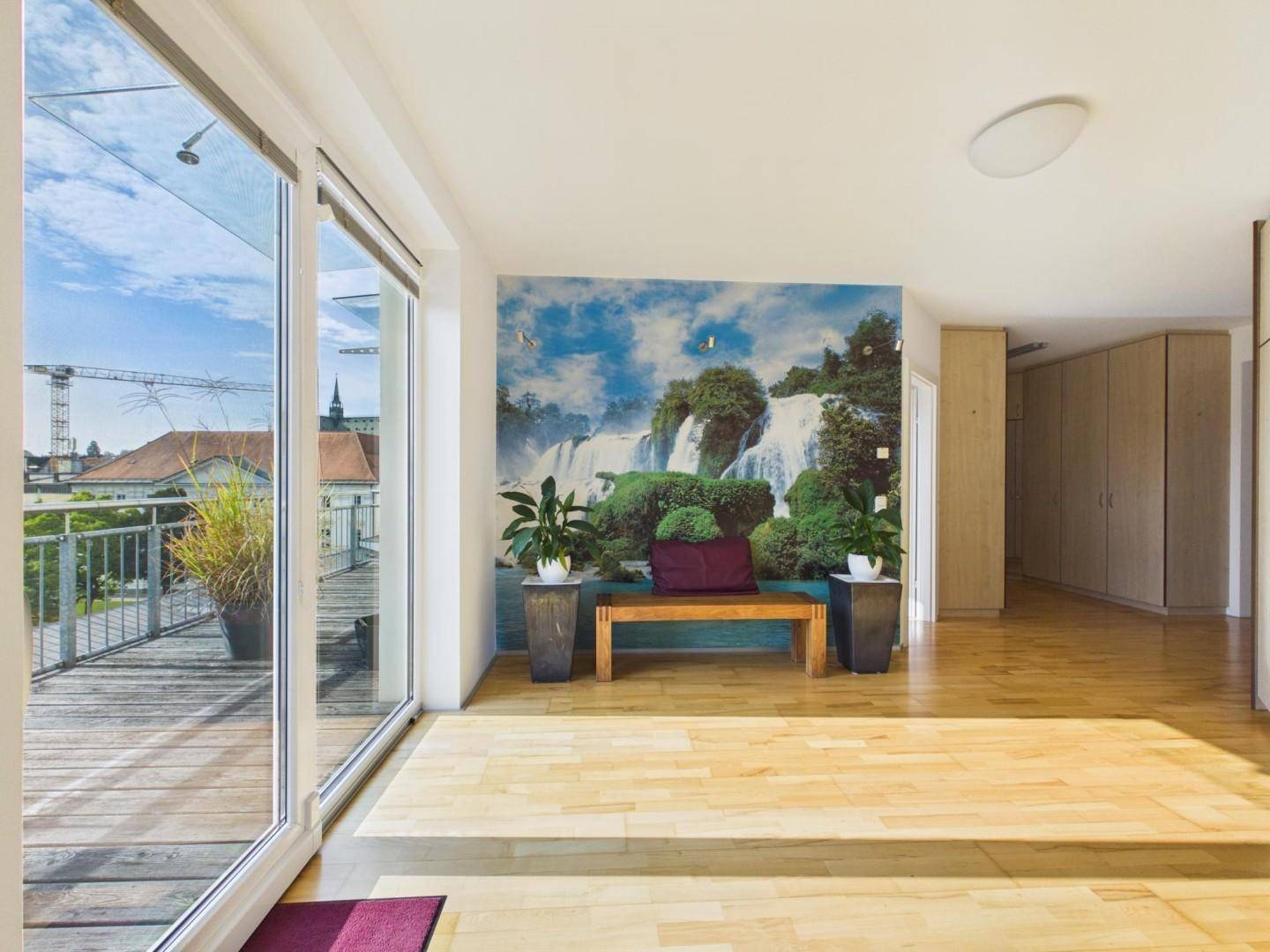 Heller Innenbereich mit Holzboden, großer Fensterfront und Zugang zu einer Holzterrasse.