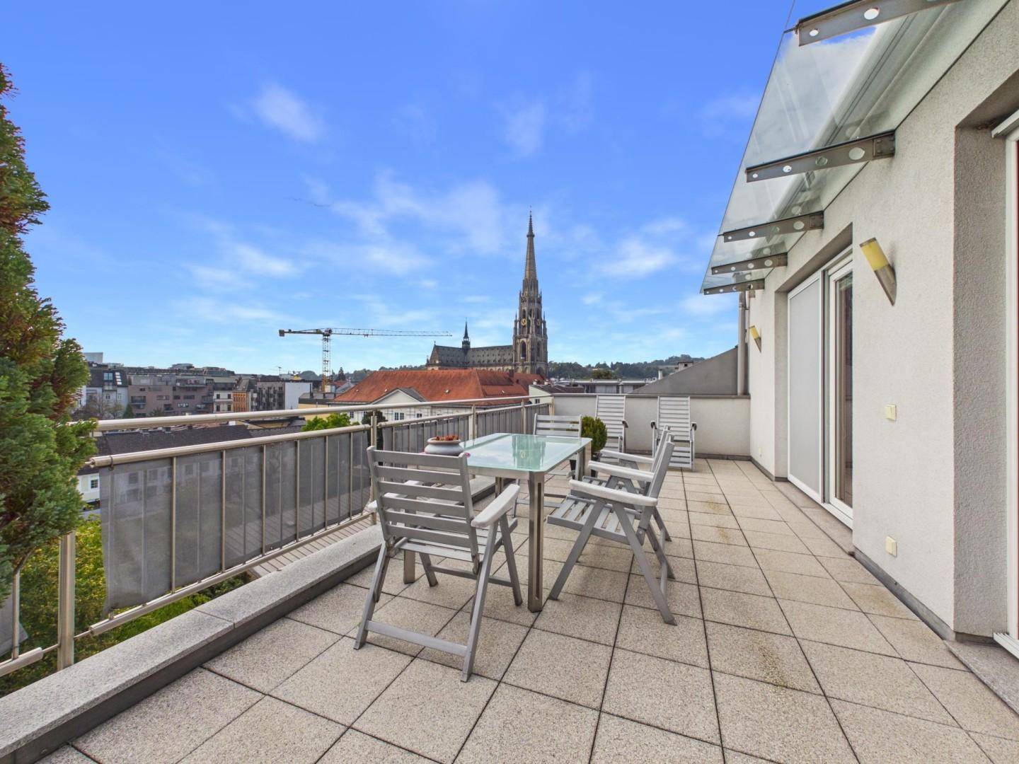 Geräumige Dachterrasse mit Sitzgelegenheiten und weitem Blick über die Stadt und eine Kirche.