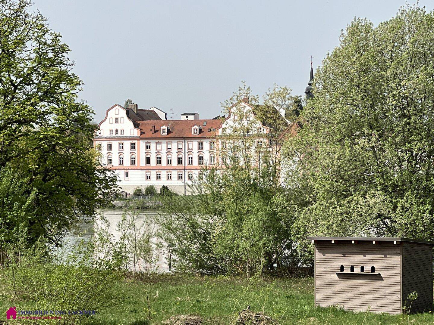 Historisches Gebäude mit rotem Dach und weißer Fassade, umgeben von Bäumen und einem Gewässer.