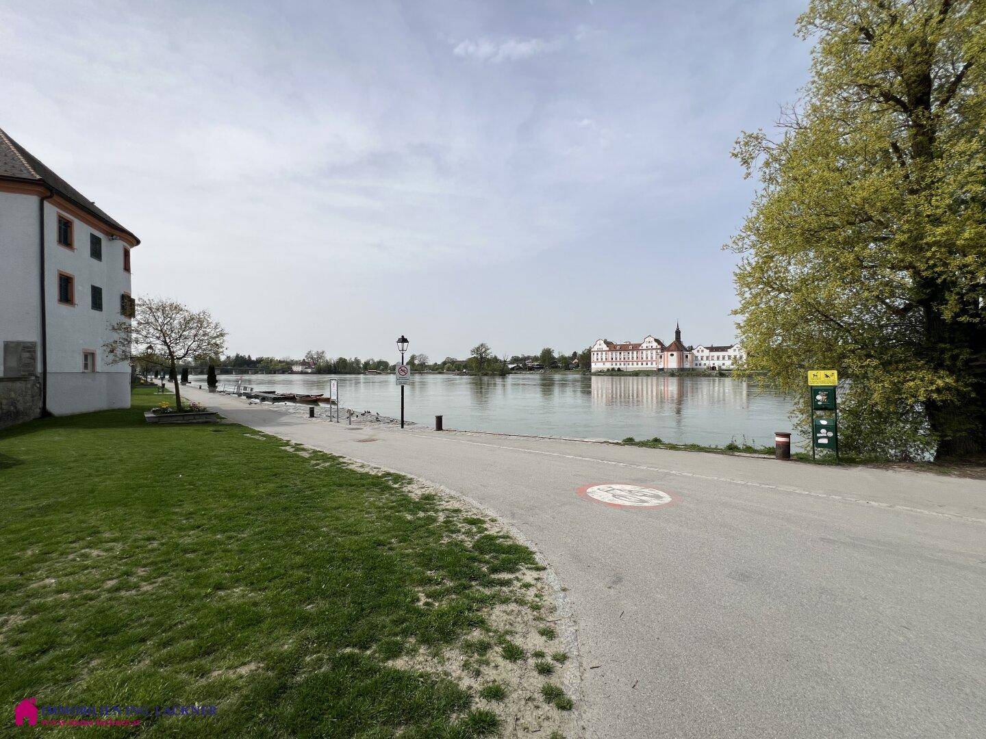 Weitläufiger Blick auf einen Fluss oder See mit Uferweg und historischem Gebäude in der Ferne.