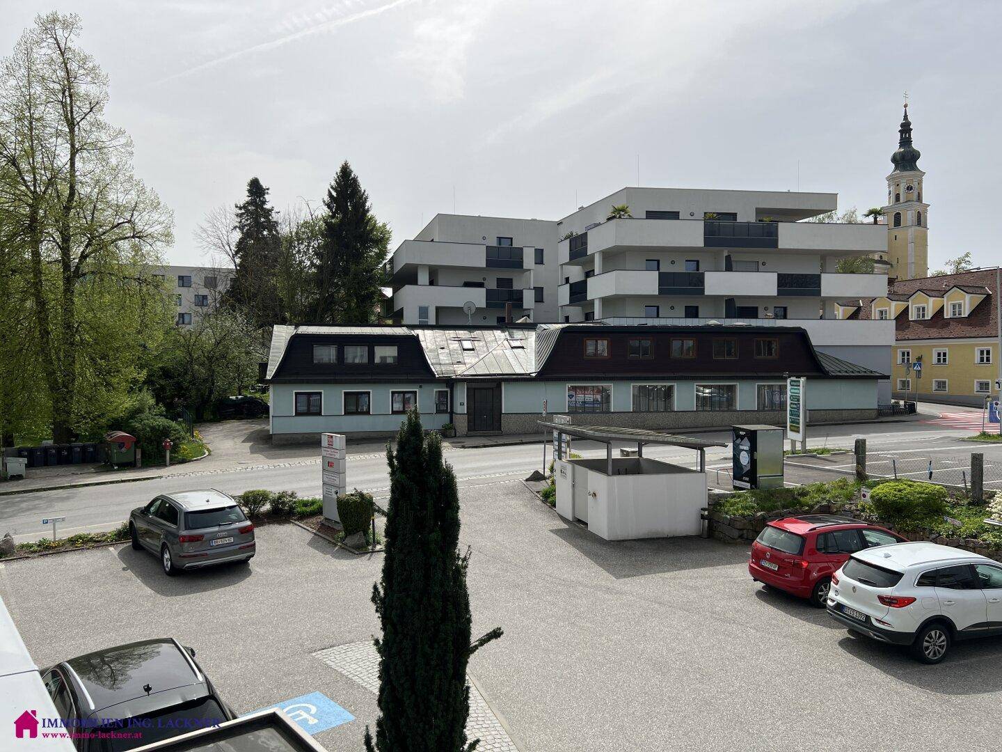 Blick auf eine städtische Szene mit verschiedenen Gebäuden, Parkplätzen und einem Kirchturm.