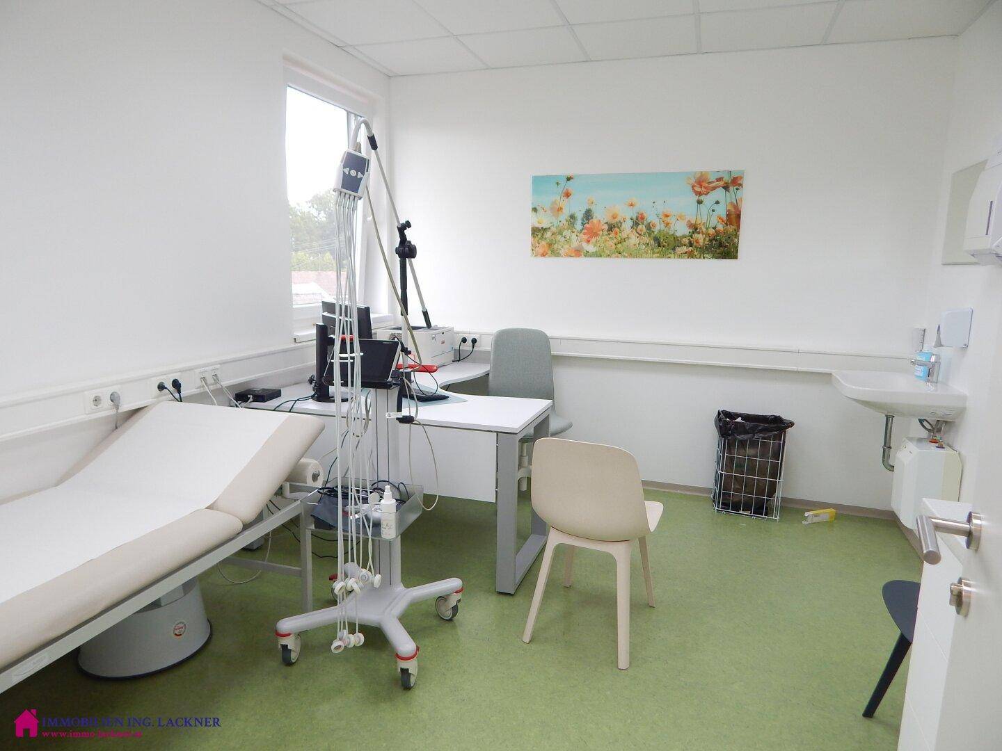 Heller Behandlungsraum mit Liege, Schreibtisch und Fenster, ausgestattet für medizinische Zwecke.