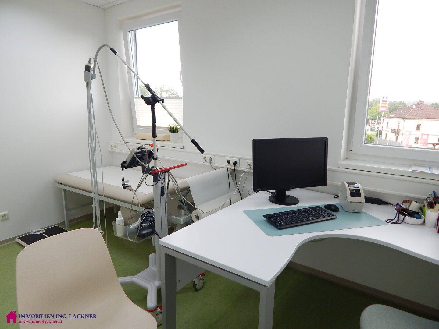 Heller Behandlungsraum mit Schreibtisch, Computer und Fenster mit Blick nach draußen.
