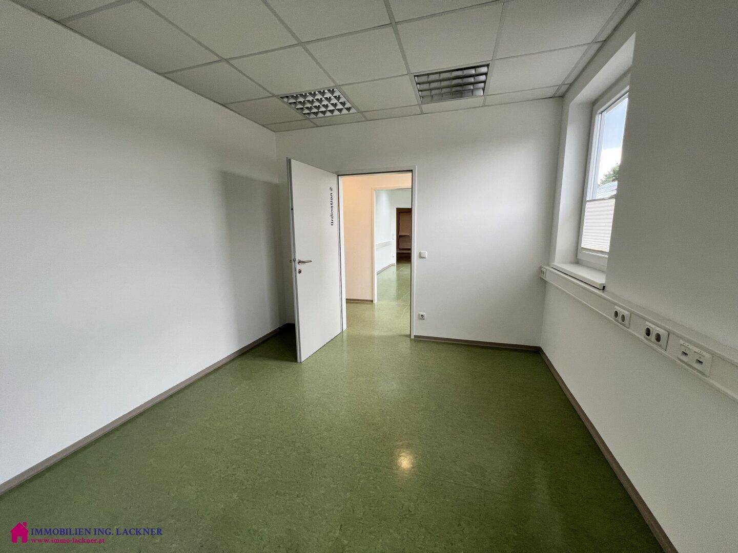 Heller Büroraum mit grünem Linoleumboden, weißen Wänden und einem Fenster mit Blick ins Grüne.