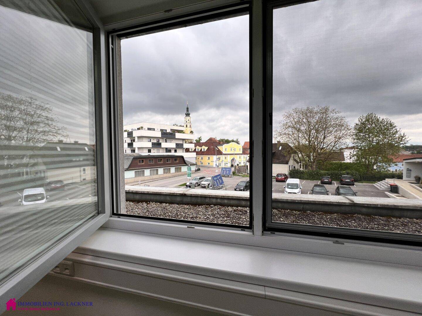 Blick aus einem Fenster auf eine städtische Umgebung mit Gebäuden, einem Kirchturm und einem Parkplatz.