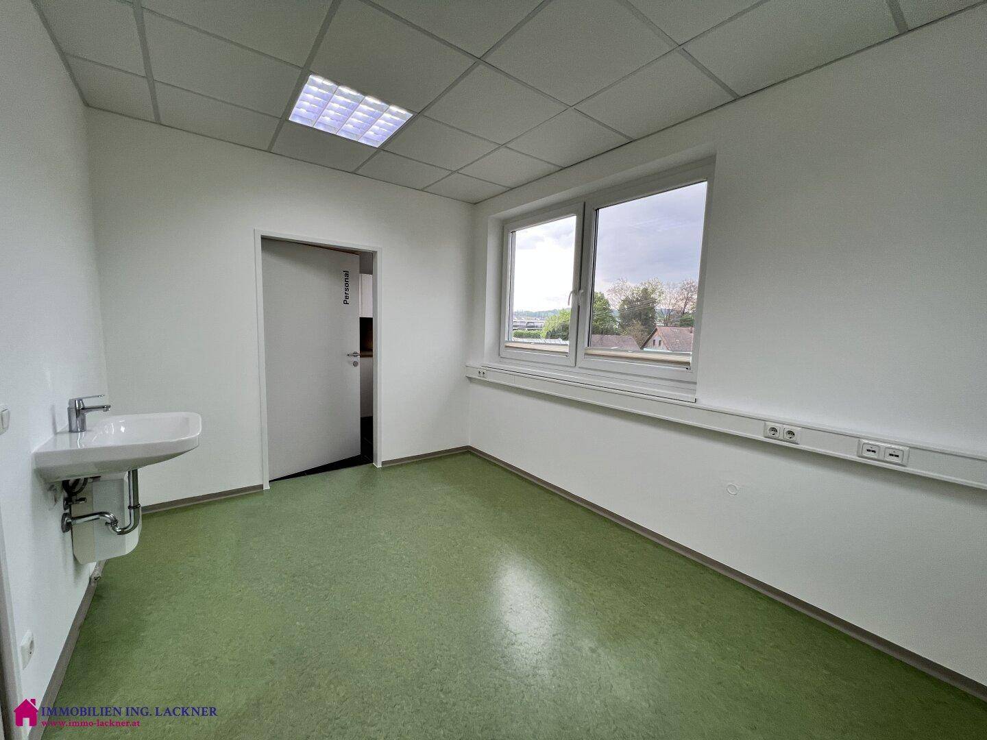 Heller Büroraum mit grünem Linoleumboden, einem Waschbecken und einem Fenster für natürliches Licht.