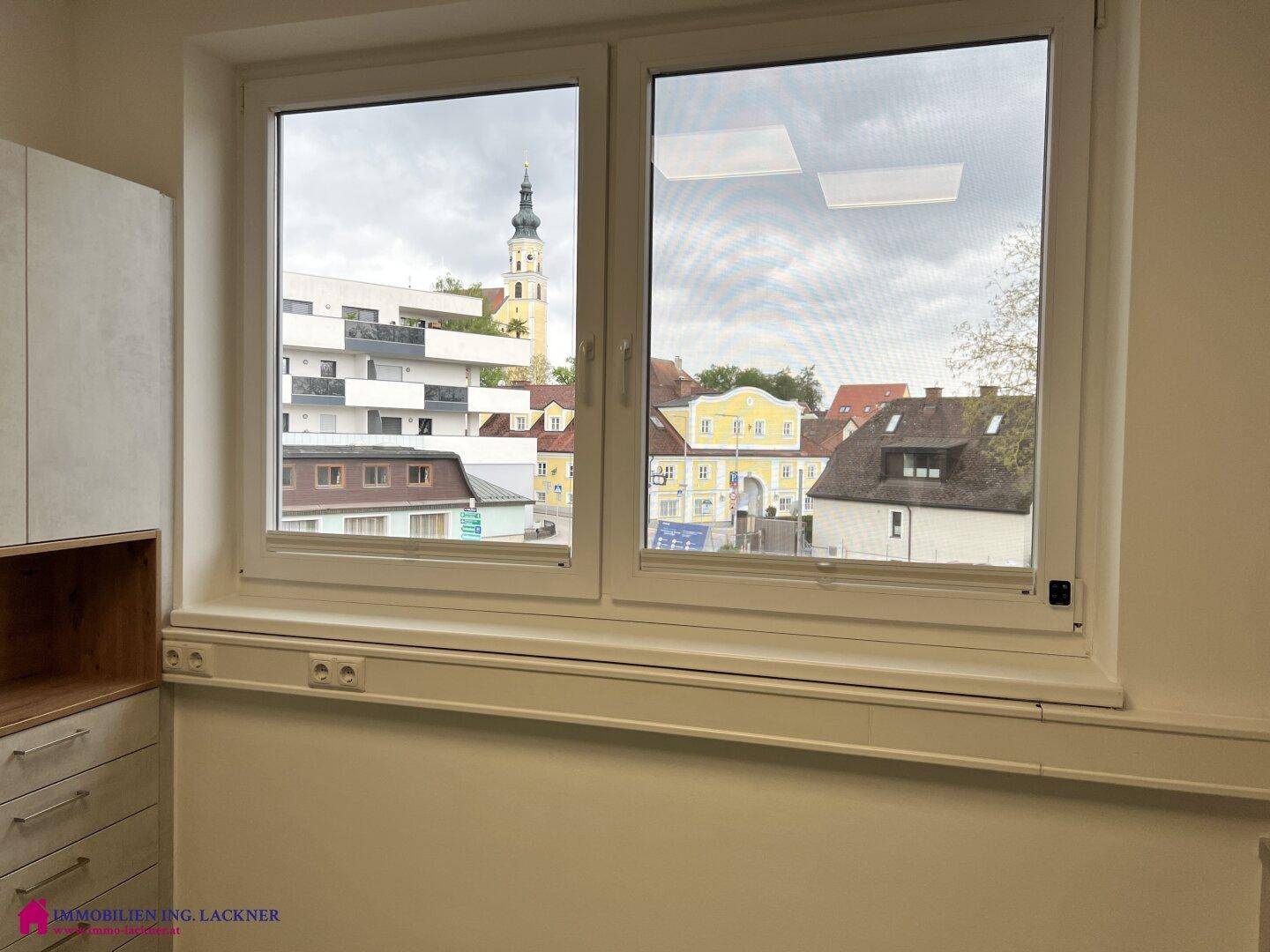 Blick aus einem Bürofenster auf eine städtische Umgebung mit einem Kirchturm und umliegenden Gebäuden.