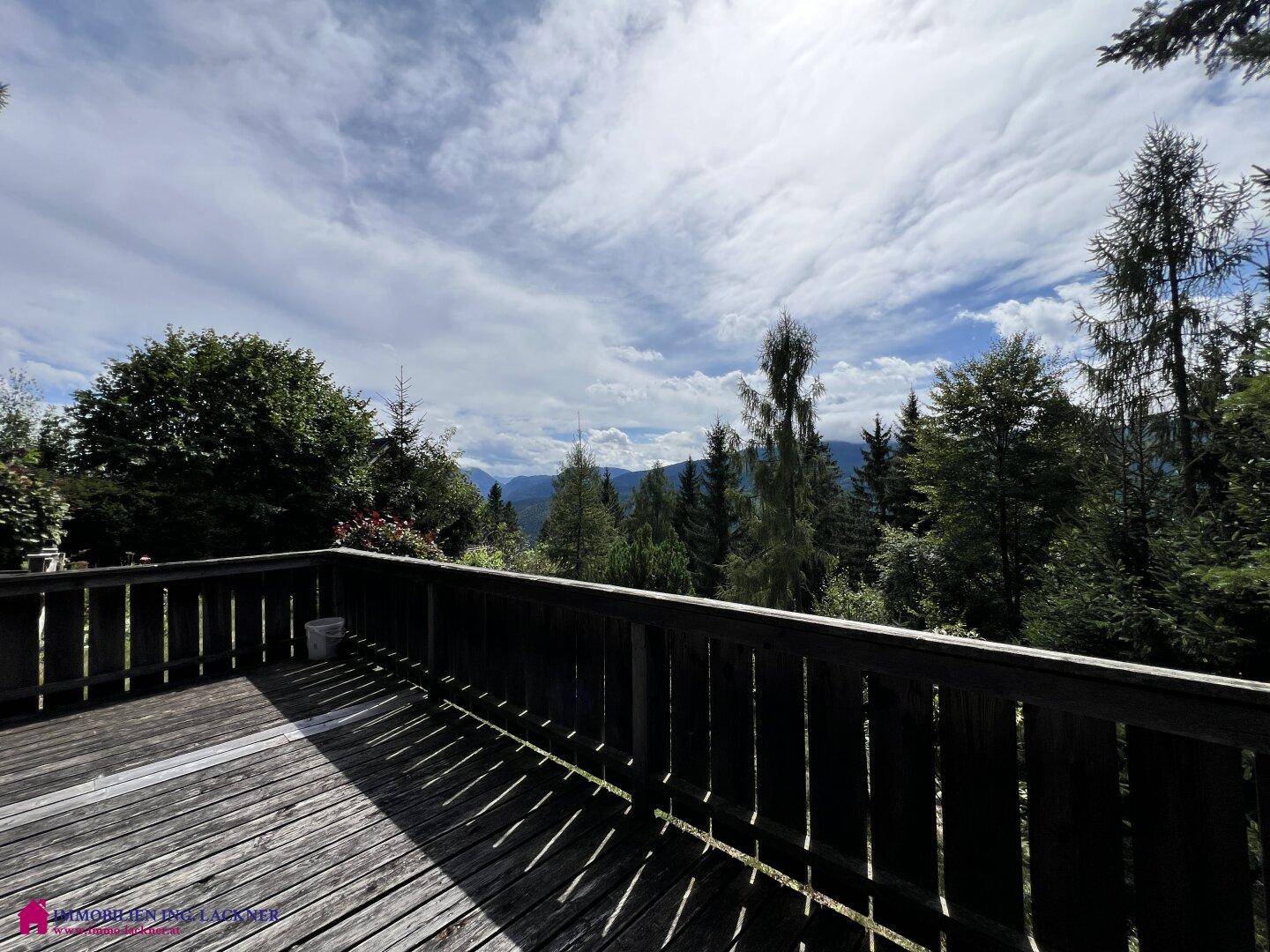 Weitblick von der Holzterrasse über dichte Wälder und majestätische Berge unter einem bewölkten Himmel.