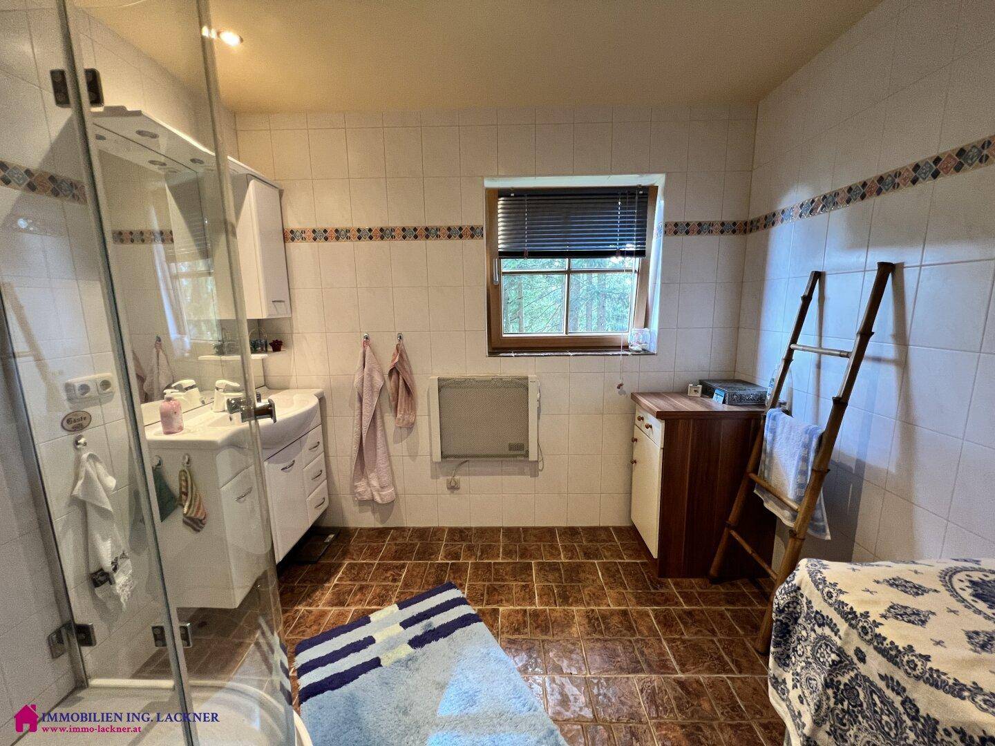 Helles Badezimmer mit Duschkabine, Waschbecken, Fenster und braunen Bodenfliesen.