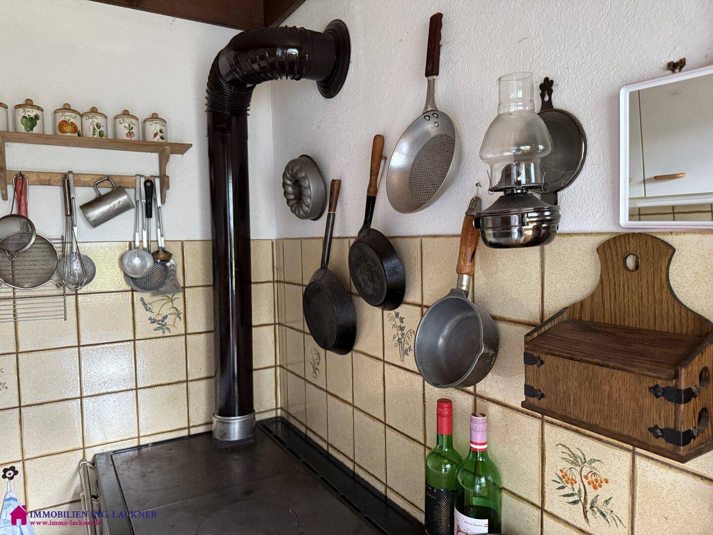 Detailansicht der Küchenwand mit rustikalen Fliesen, hängenden Kochutensilien und dem Ofenrohr des Holzherdes.