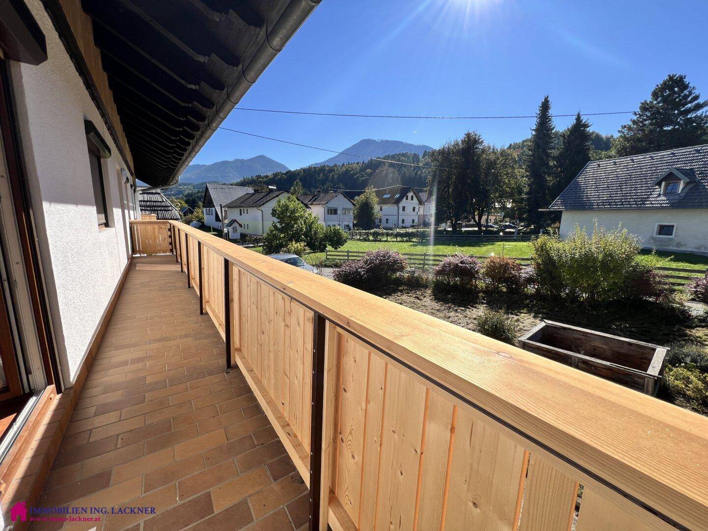 Langer Balkon mit Holzgeländer und Fliesenboden, bietet einen schönen Blick auf die Berge und Kirche.