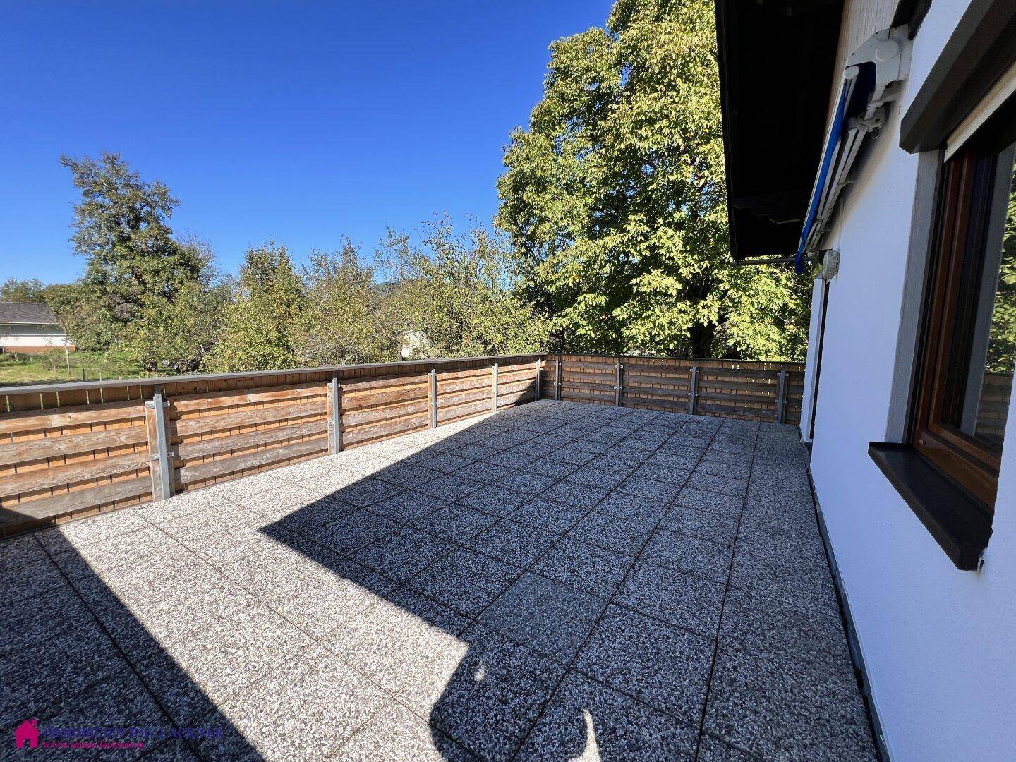 Große Terrasse mit Fliesenboden und Holzzaun, angrenzend an die Hausfassade mit Fenster.