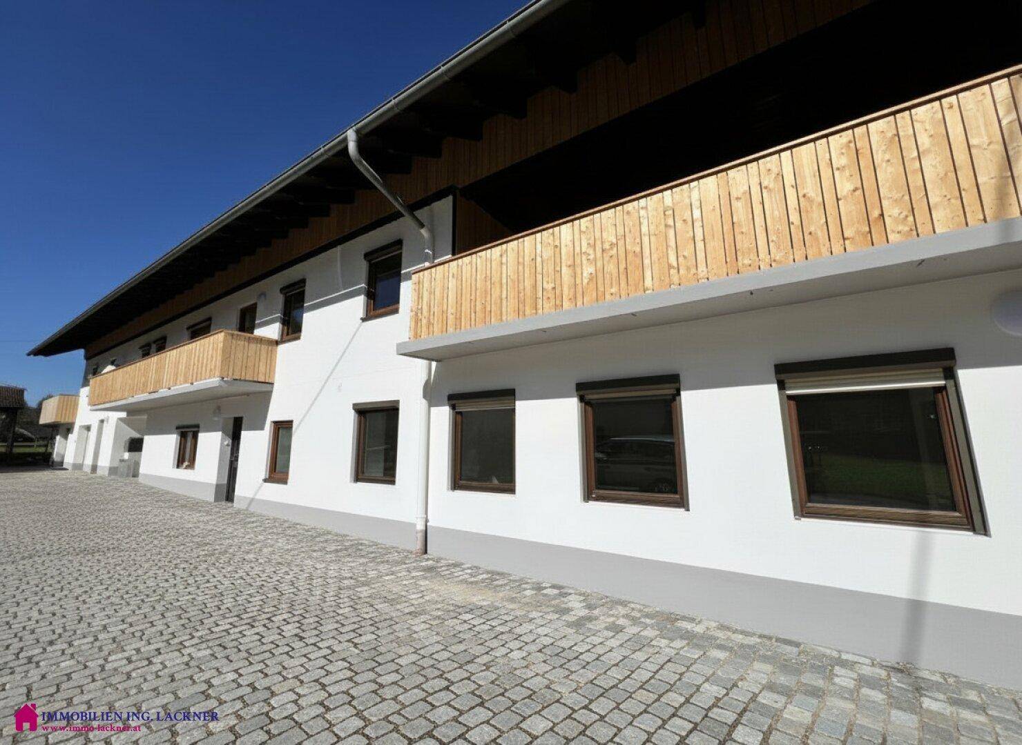 Seitenansicht des Gebäudes mit mehreren Fenstern, Rollläden und Balkonen, gepflasterter Vorplatz.