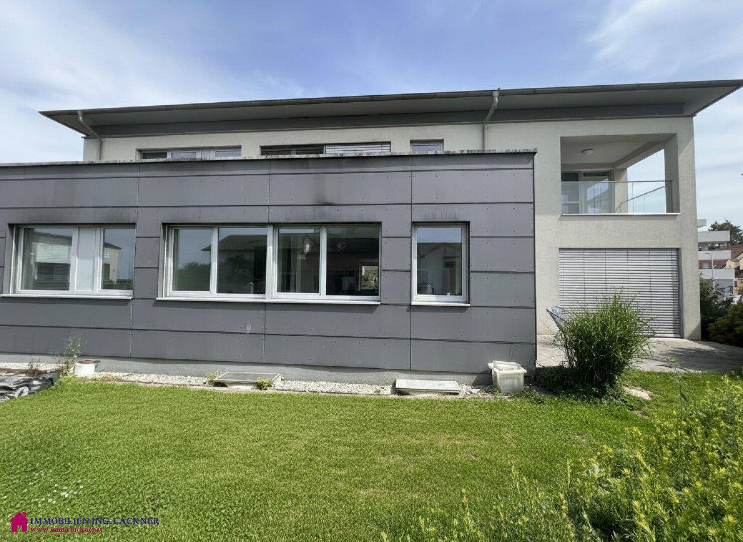 Rückseite des modernen Gebäudes mit gepflegtem Rasen und einer überdachten Loggia im Obergeschoss.