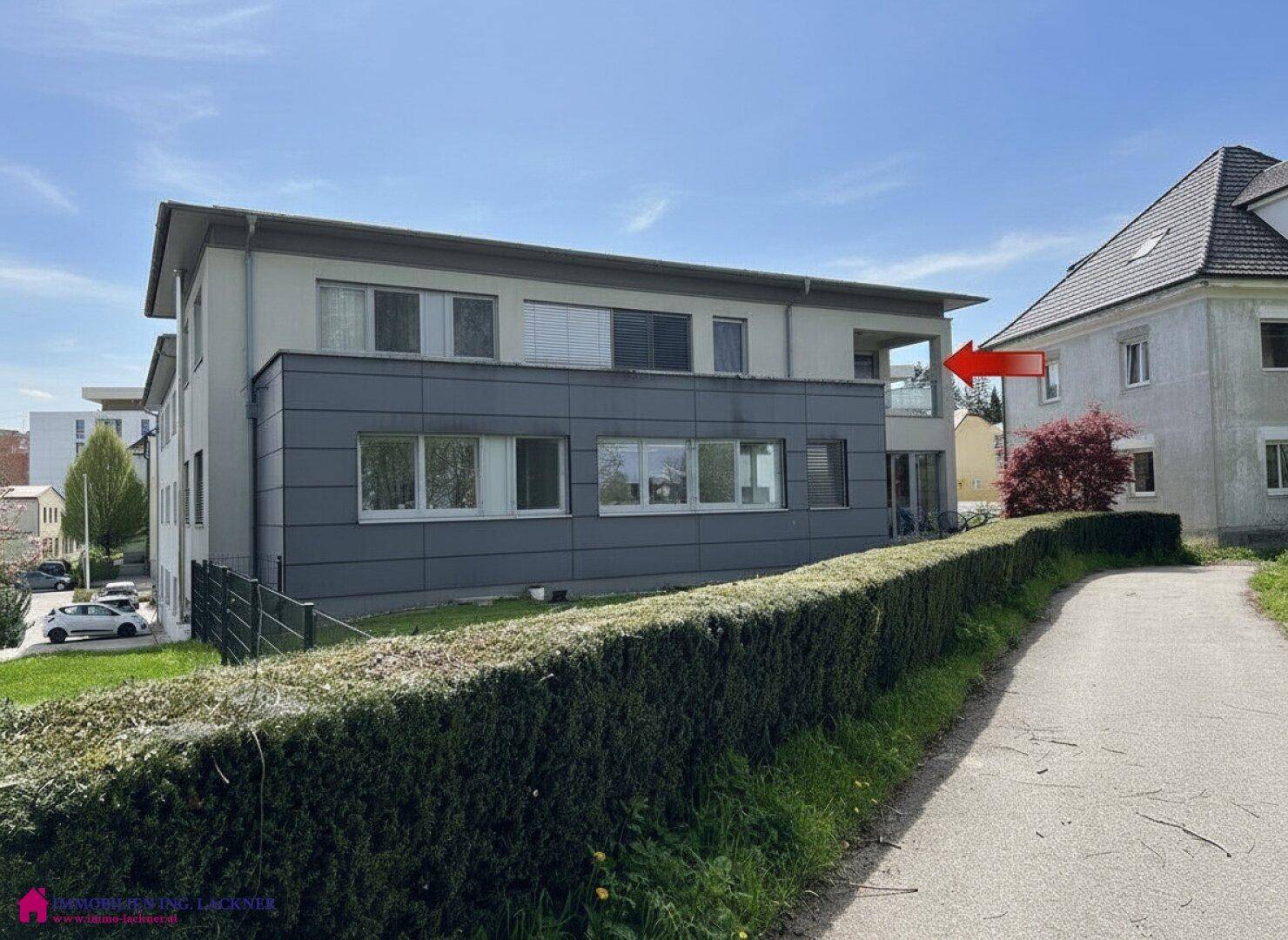 Rückansicht eines modernen Gebäudes mit grauer Verkleidung und einem Balkon im oberen Stockwerk, umgeben von Hecken.