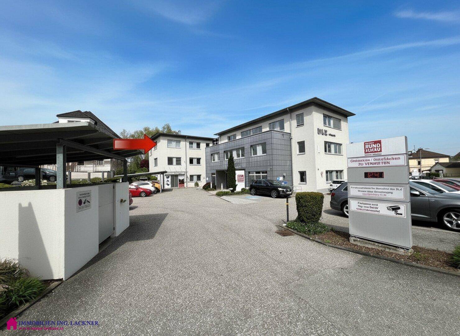 Gebäudekomplex mit großem Parkplatz und Carport, umgeben von Grünflächen und weiteren Gebäuden.