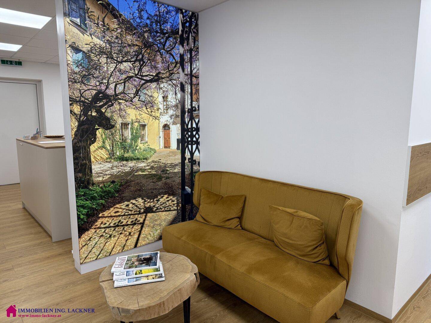 Gemütlicher Wartebereich mit einem Sofa und einer großformatigen Wandtapete, die einen Innenhof darstellt.
