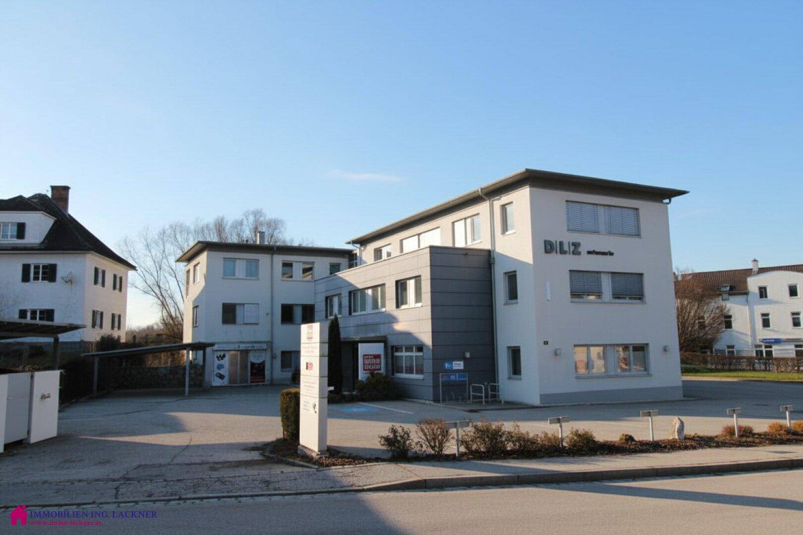Modernes Geschäftsgebäude mit großem Parkplatz und klarem Himmel, umgeben von weiteren Gebäuden.