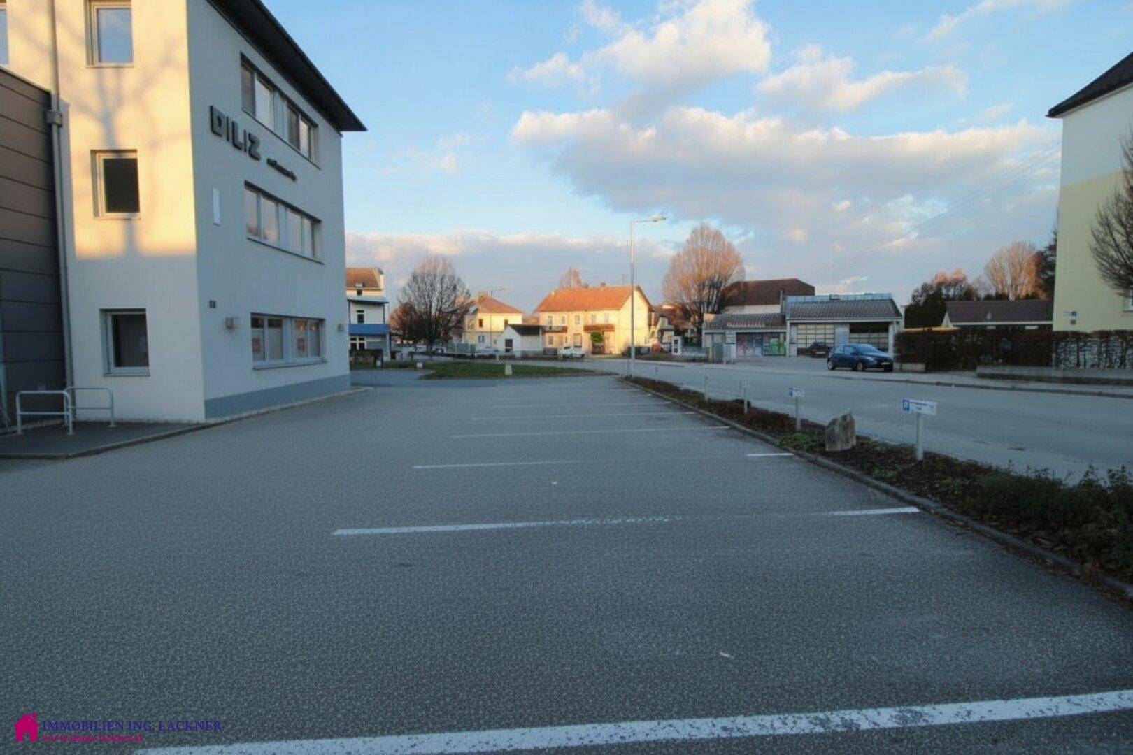 Großer, leerer Parkplatz vor dem Geschäftsgebäude mit Blick auf die angrenzende Straße und Wohnhäuser.