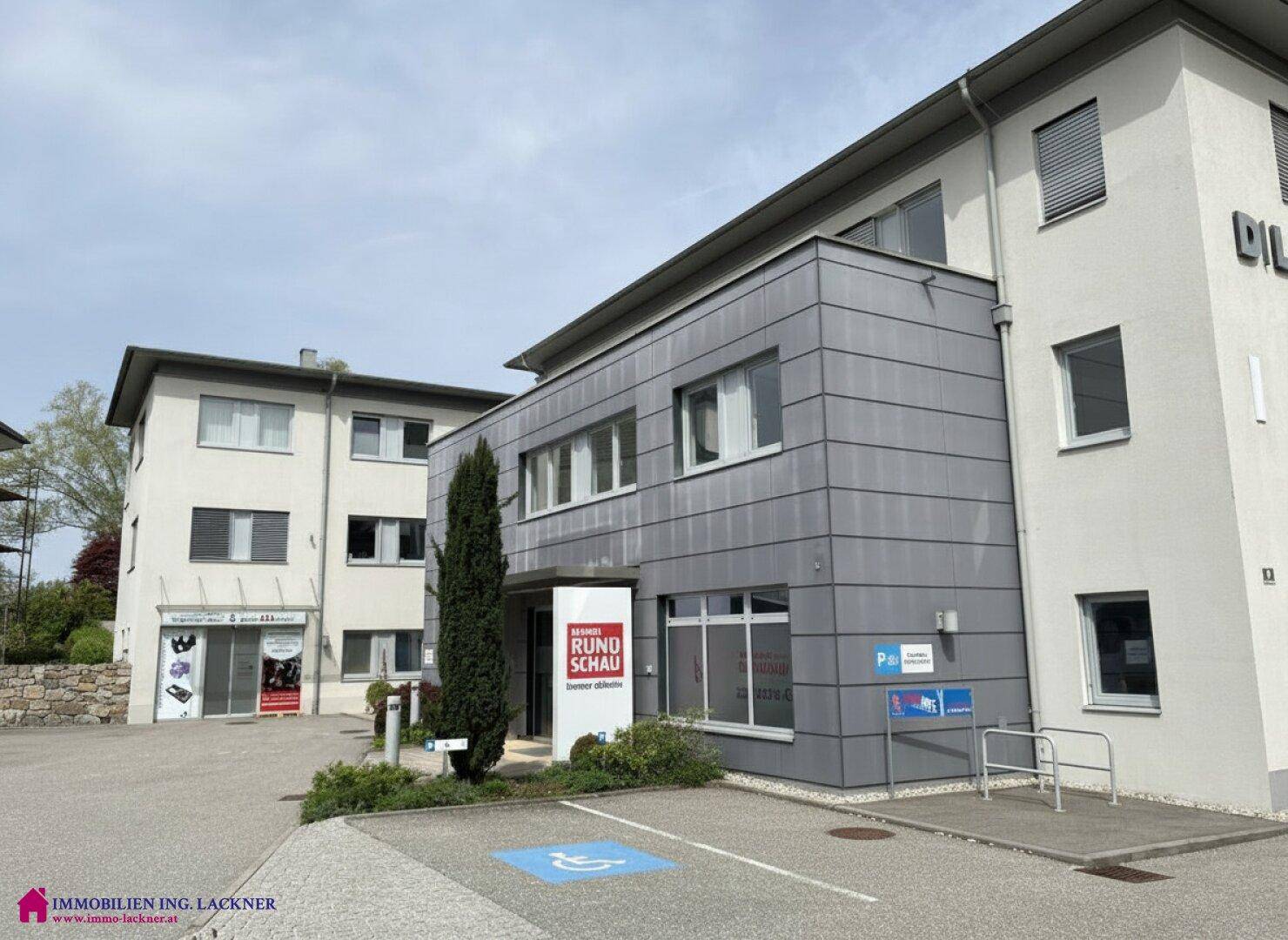 Modernes Geschäftsgebäude mit grauen Fassadenelementen und großen Fenstern, umgeben von Grün.