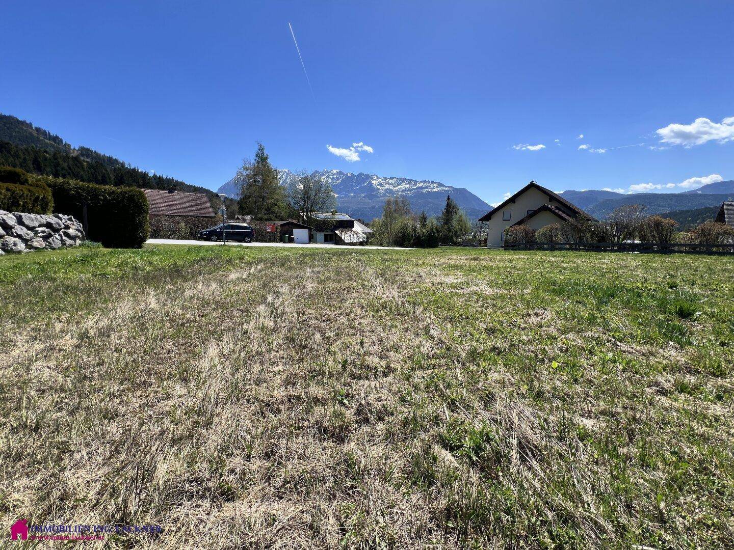 Offenes Wiesengrundstück mit Häusern im Hintergrund und klarer Sicht auf die Alpen.
