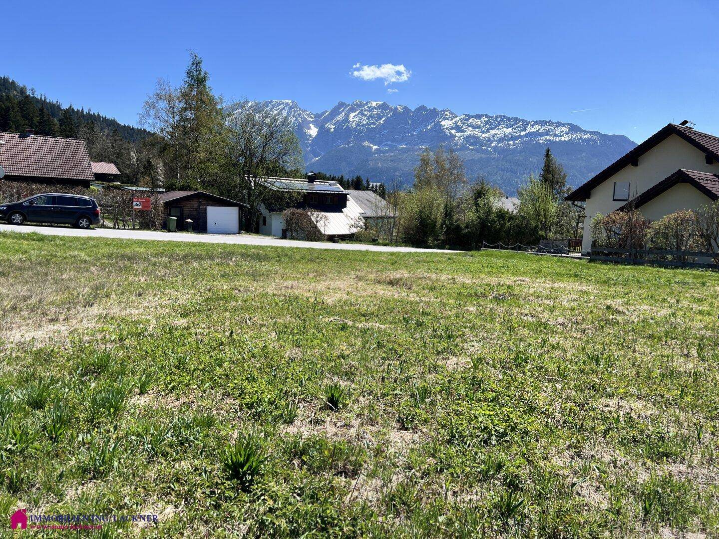Weitläufige Grünfläche mit Häusern im Hintergrund und einem weiten Blick auf die Alpen.