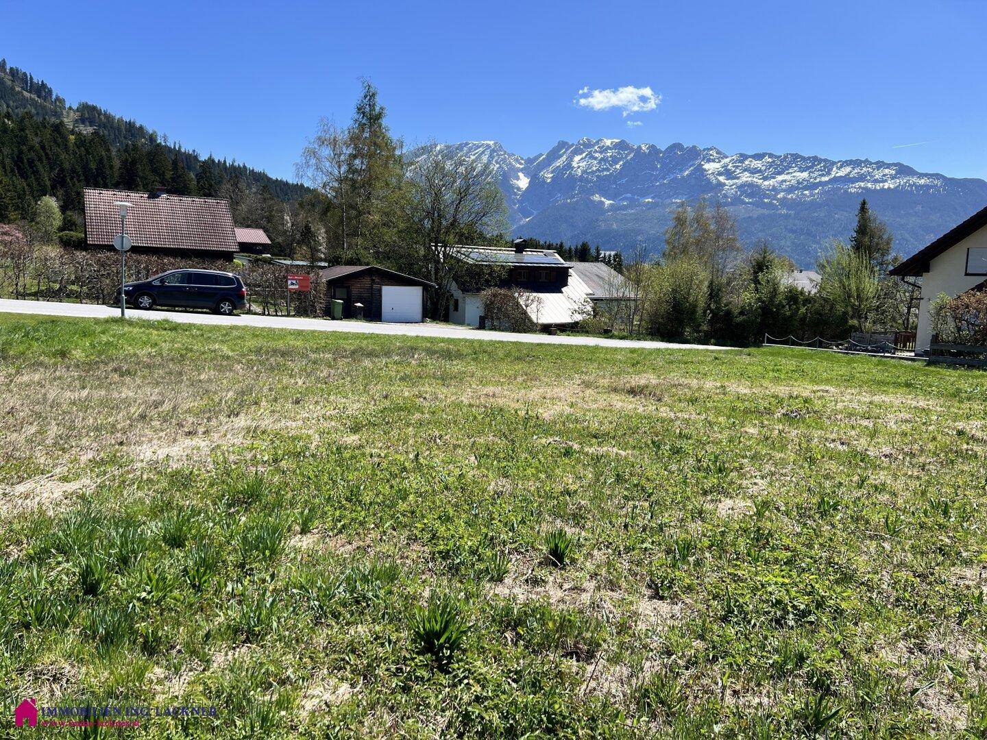 Offenes Grundstück mit grüner Wiese, umgeben von ländlicher Bebauung und Bergen.