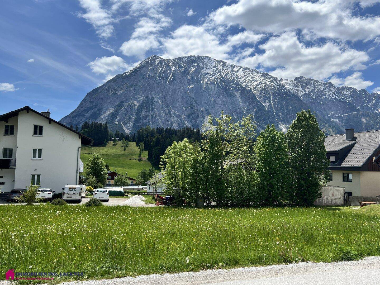 Sonnige Wiese mit Blick auf Wohngebäude und die imposante, schneebedeckte Berglandschaft.