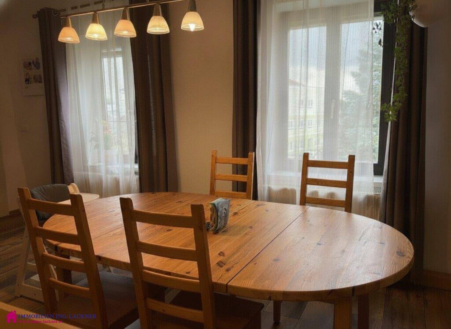 Heller Essbereich mit großem Holztisch, Holzstühlen und Fenstern mit Vorhängen.