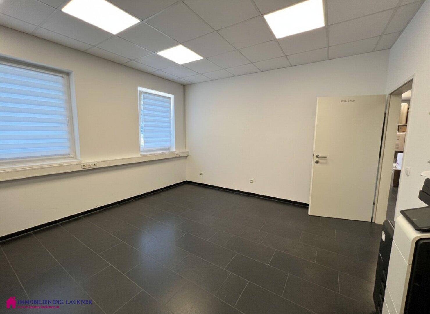 Helles Büro mit dunklem Fliesenboden, zwei Fenstern und einer offenen Tür, die zu weiteren Bereichen führt.