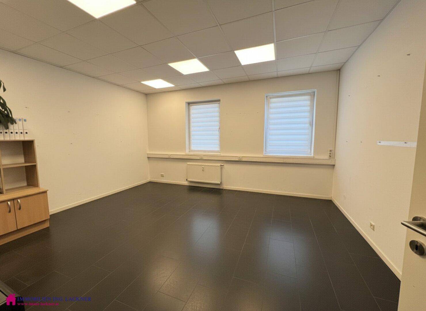Geräumiges Büro mit dunklem Fliesenboden, zwei Fenstern und einem kleinen Holzschrank.
