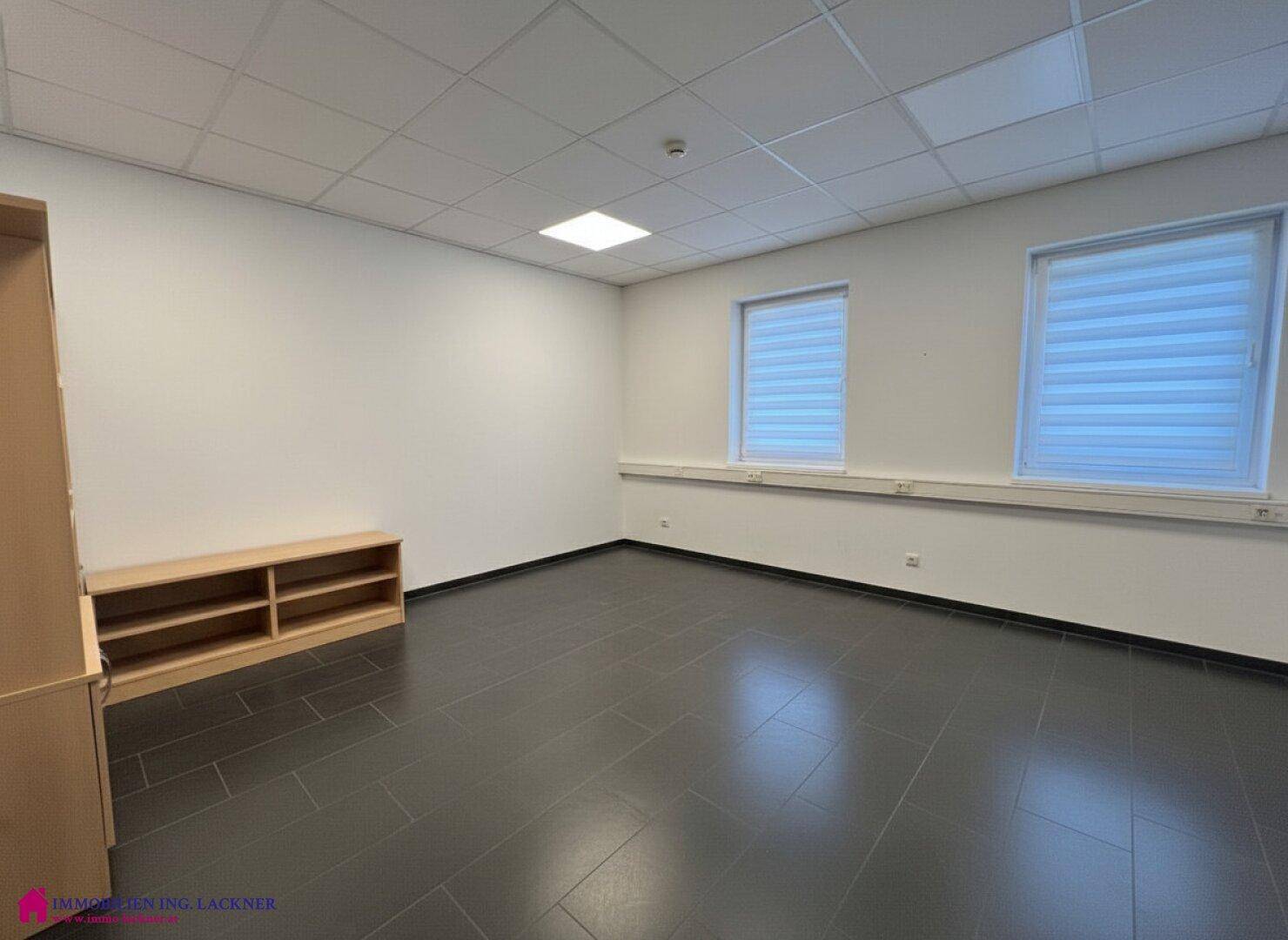 Leeres Büro mit dunklem Fliesenboden, zwei Fenstern und einem niedrigen Holzregal.