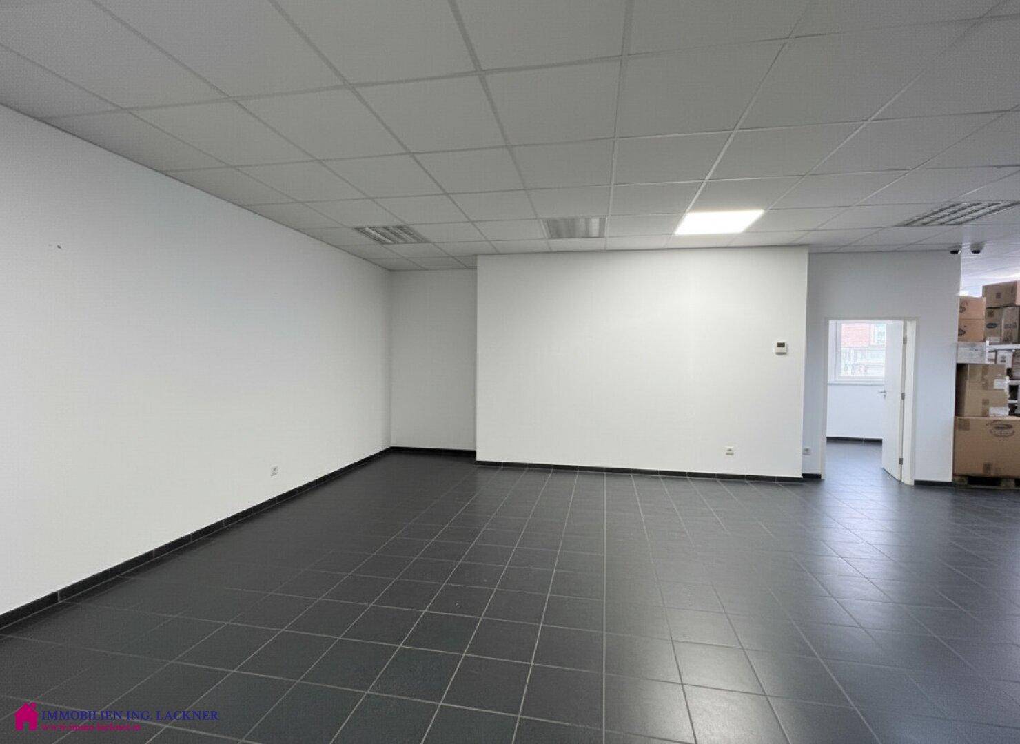 Geräumiger Bürobereich mit dunklem Fliesenboden und weiß gestrichenen Wänden, angrenzend an einen Lagerbereich.
