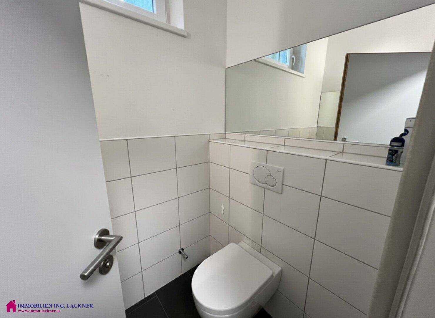 Modernes WC mit weißen Fliesen, wandhängender Toilette und großem Spiegel.