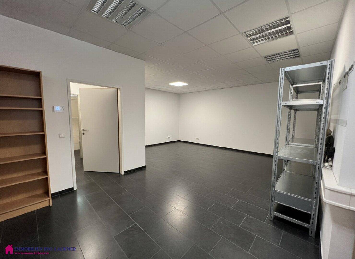 Geräumiger Bürobereich mit dunklem Fliesenboden, Holzregal und Metallregalen, ideal für flexible Nutzung.