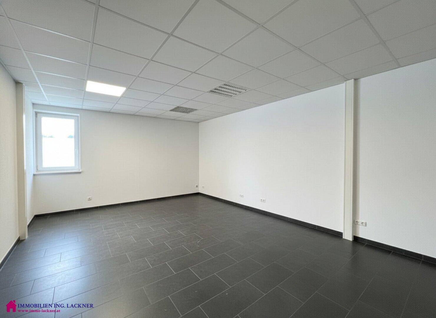 Helles, leeres Büro mit dunklem Fliesenboden und einem Fenster, das viel Tageslicht hereinlässt.