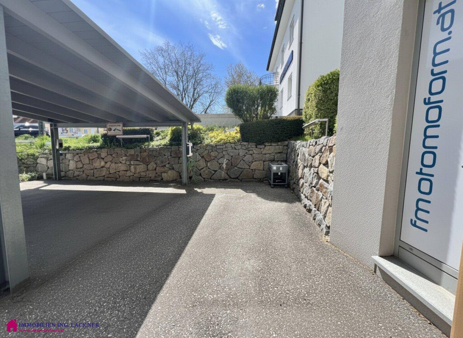 Überdachter Parkplatz mit Natursteinmauer und Ladestation für Elektrofahrzeuge, Zufahrt sichtbar.