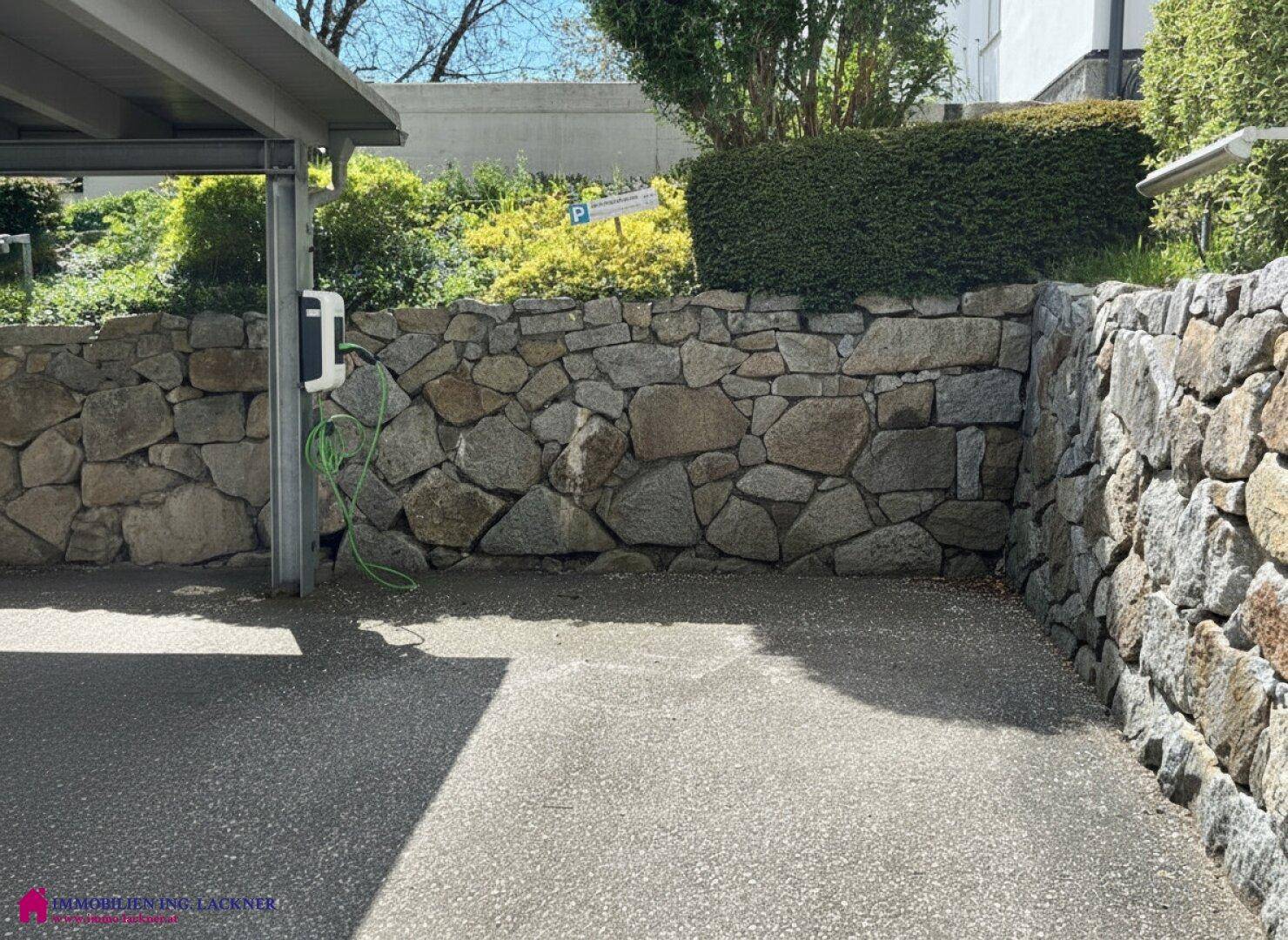 Überdachter Parkplatz mit Natursteinmauer und einer Ladestation für Elektrofahrzeuge.