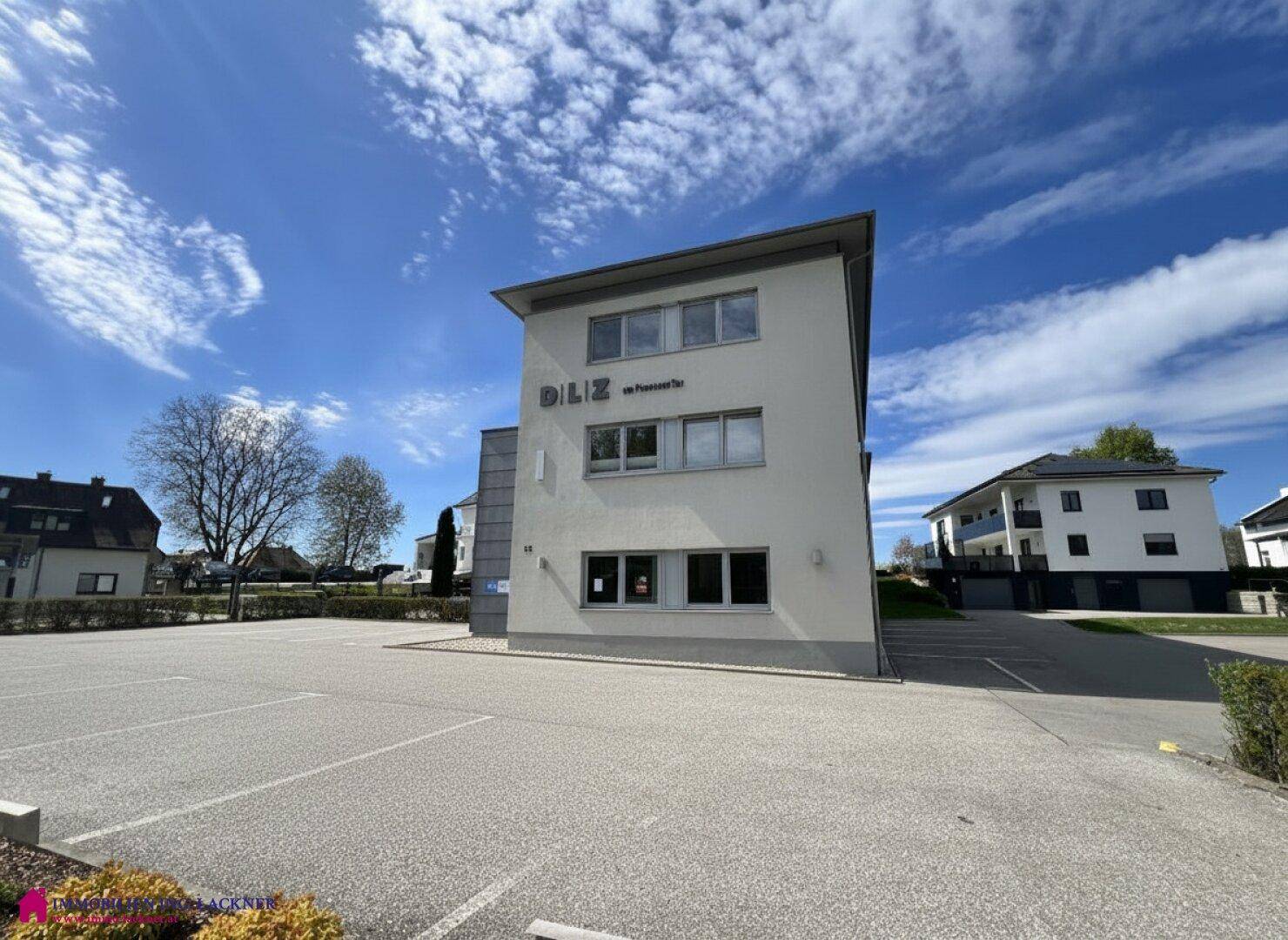 Modernes Gebäude mit heller Fassade und großem Parkplatz unter blauem Himmel.