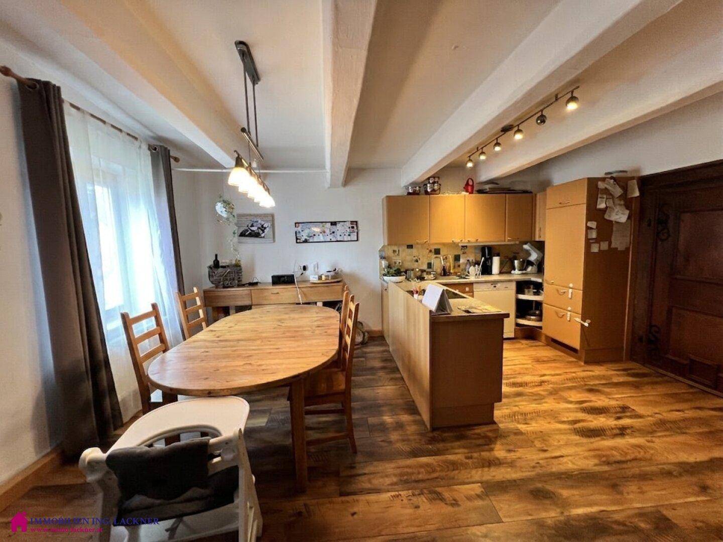Heller Essbereich mit Holztisch und offener Küche unter sichtbaren Deckenbalken.