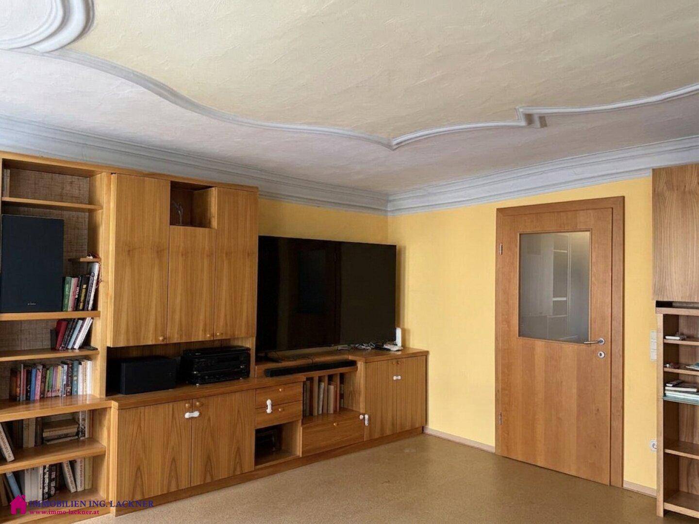 Wohnzimmer mit großer Holzschrankwand, Fernseher und dekorativer Stuckdecke.