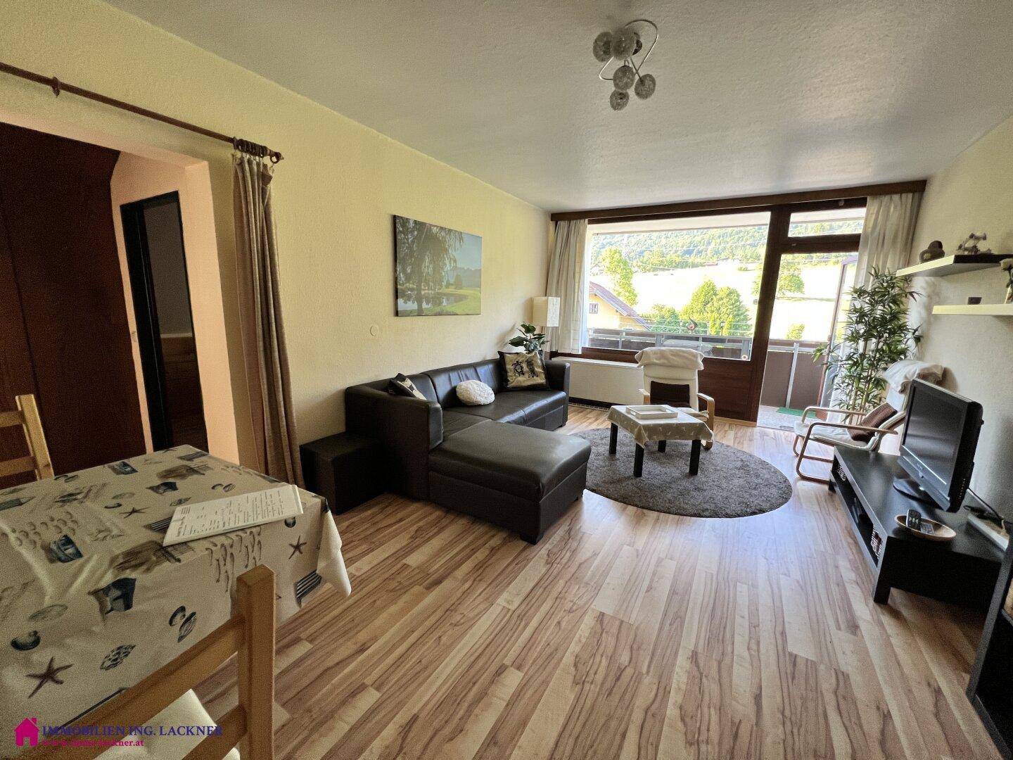 Gemütlicher Wohn-Essbereich mit Laminatboden, dunklem Sofa und Zugang zu einem Balkon mit Bergblick.