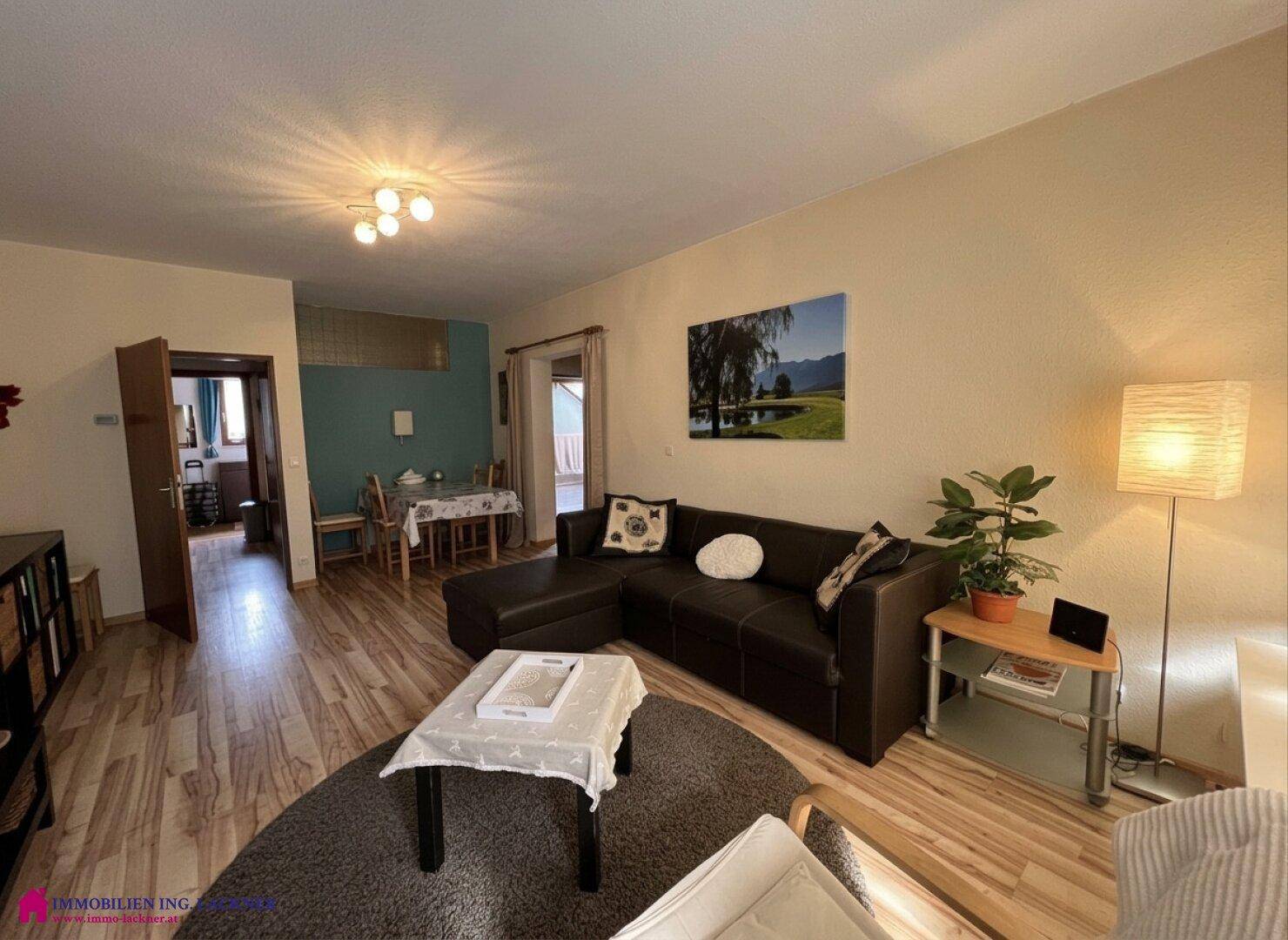 Heller Wohnbereich mit beigem Sofa, Laminatboden und einer Stehlampe, die warmes Licht spendet.
