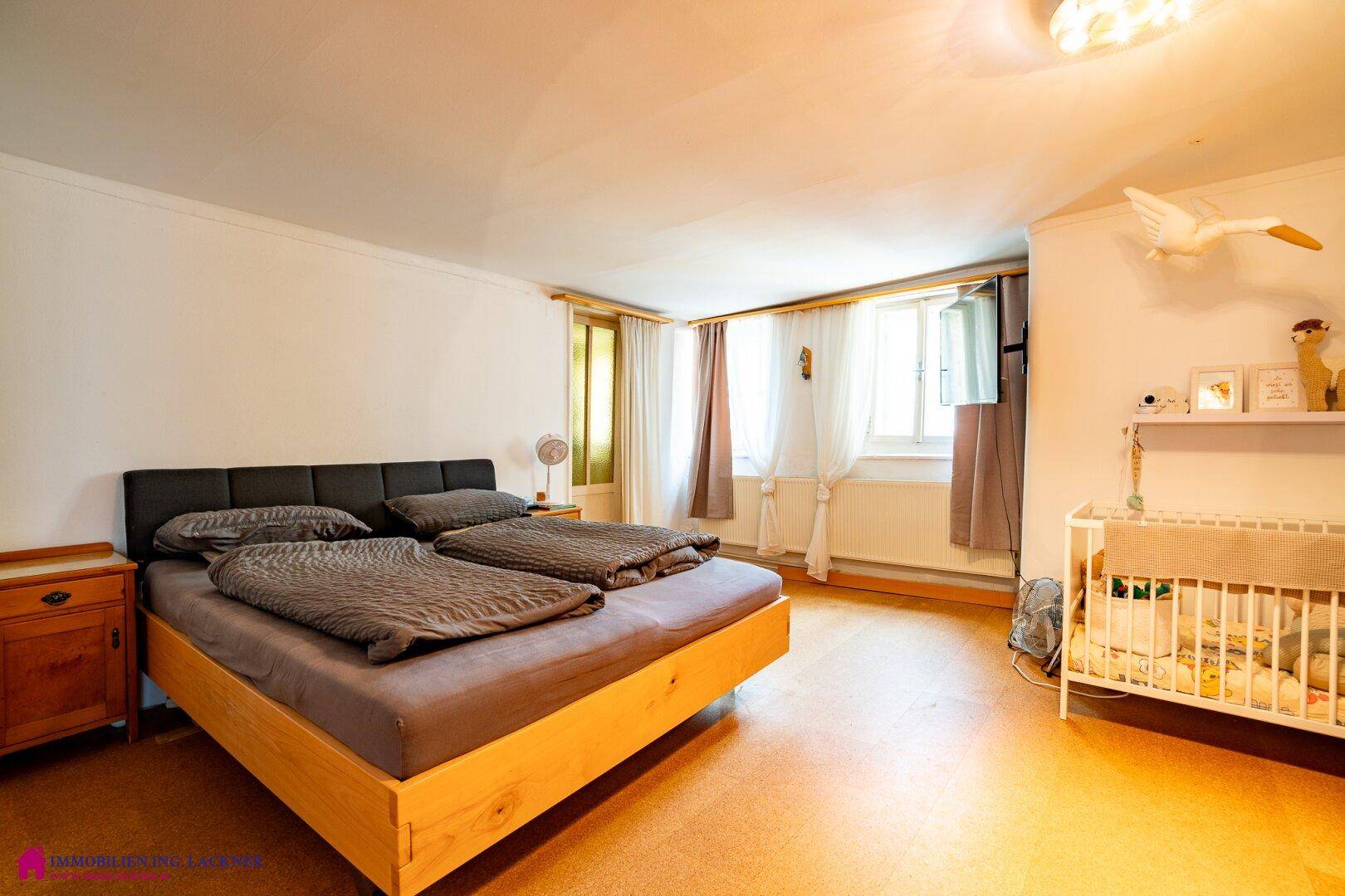 Helles Schlafzimmer mit Doppelbett, Babybett und Fenstern mit Vorhängen.