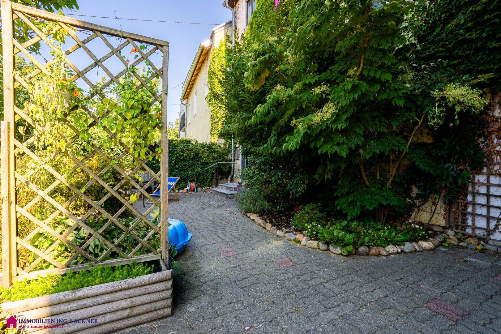 Gepflasterter Weg führt durch einen üppigen Gartenbereich mit Holzspalier und dichter Bepflanzung.