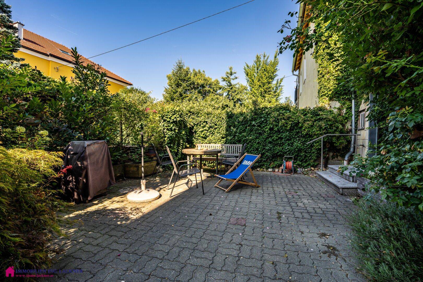 Sonniger Terrassenbereich mit Gartenmöbeln, umgeben von hohen Hecken und Bäumen, ideal zum Entspannen.